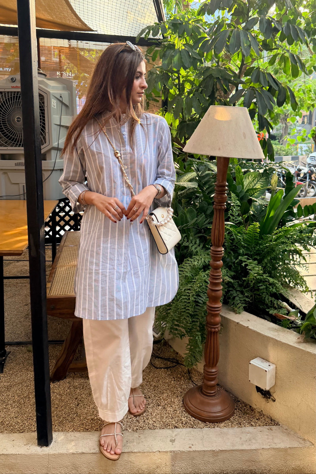 Woman standing outdoors in a casual setting with plants and a lamp.