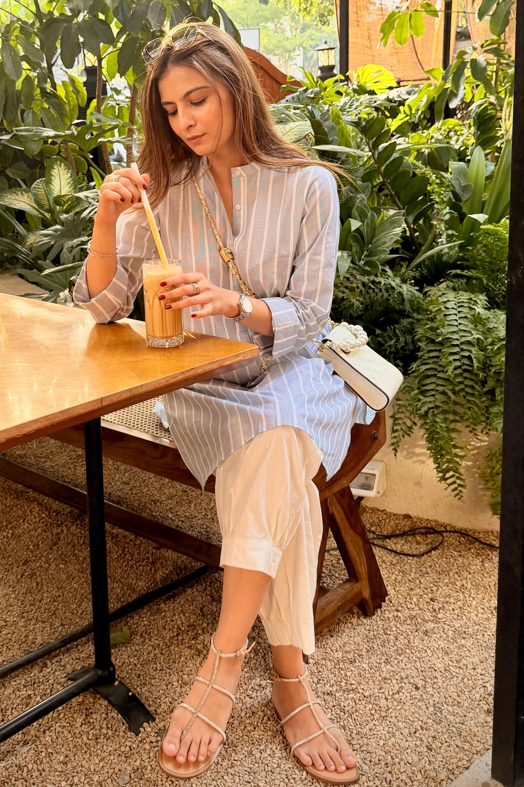 Woman sitting at a table outdoors, holding a drink, surrounded by greenery.